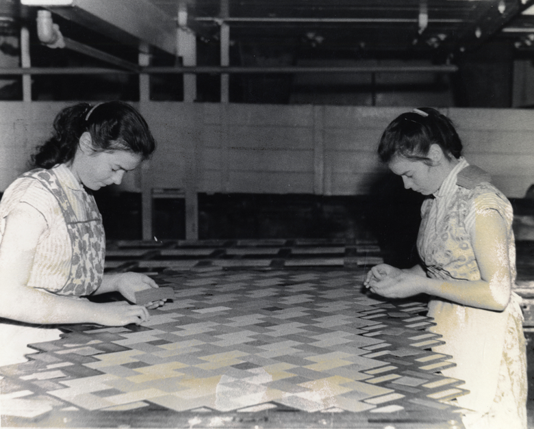 Two women laying linoleum tiles to make patterned linoleum. 