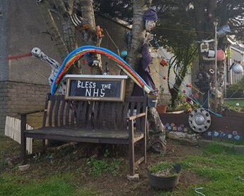 Photo of a wooden bench with Bless the NHS sign attached to it. Various other decorations, including the rainbow, are surrounding the sign.- 