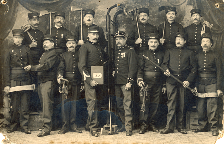 A black and white photograph showing a group of men posing in an interior. Each man wears a dark uniform and a peak cap, and several have large moustaches. Some of them carry objects associated with fire-fighting, including a hose and a ladder.