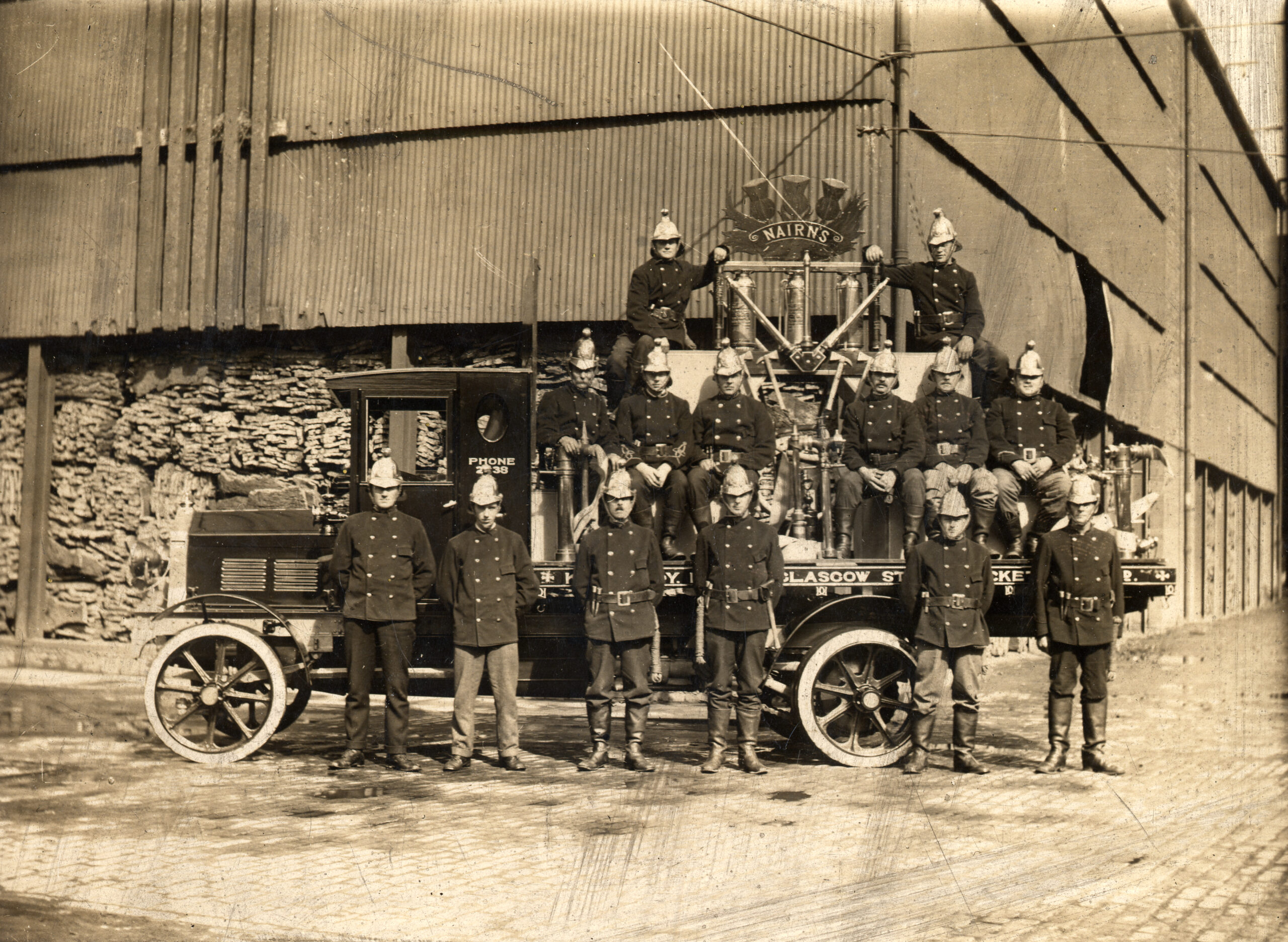 A black and white photograph of a group of men standing on a cart outside a factory. The men all wear dark uniforms, and dome-shaped helmets. A sign in the shape of a wooden thistle is balanced on top of the cart behind them.