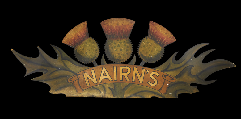 A painted wooden sign in the shape of three thistles. The thistles are green with fluffy pinkish purple petals. Across the centre of the logo is a curved yellow ribbon with 'Nairn's' printed on it in capital letters.